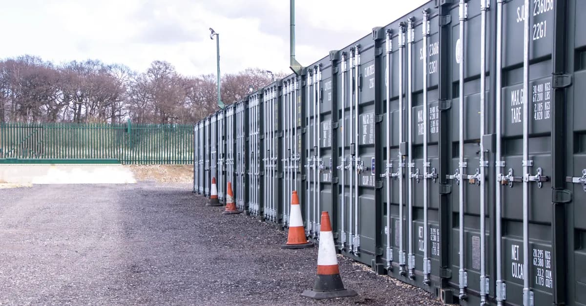 Row of grey shipping container storage units beside a gravel yard, with traffic cones along the doors
